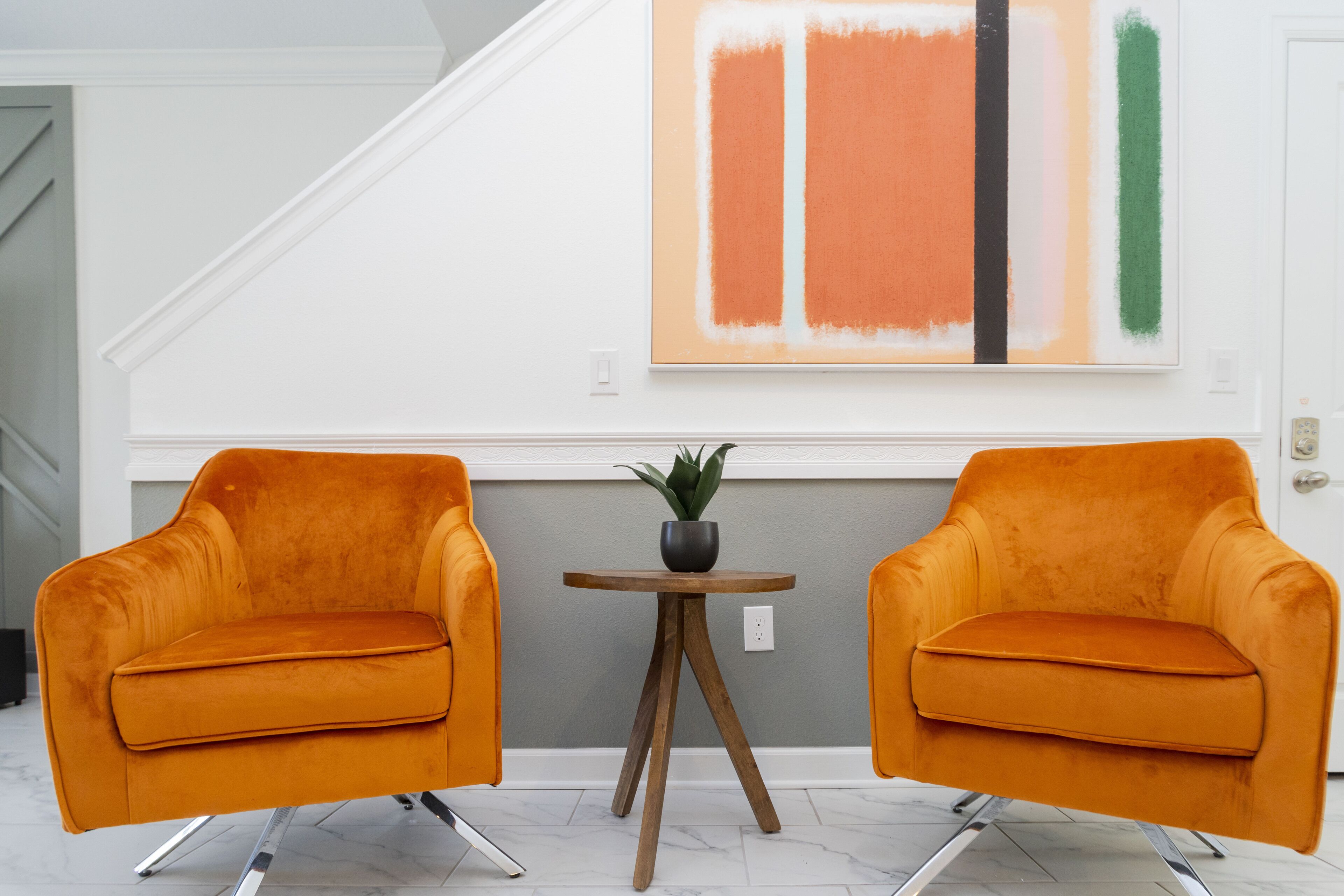 Two orange chairs with a small table and a plant between them, set against a wall with a large abstract painting.