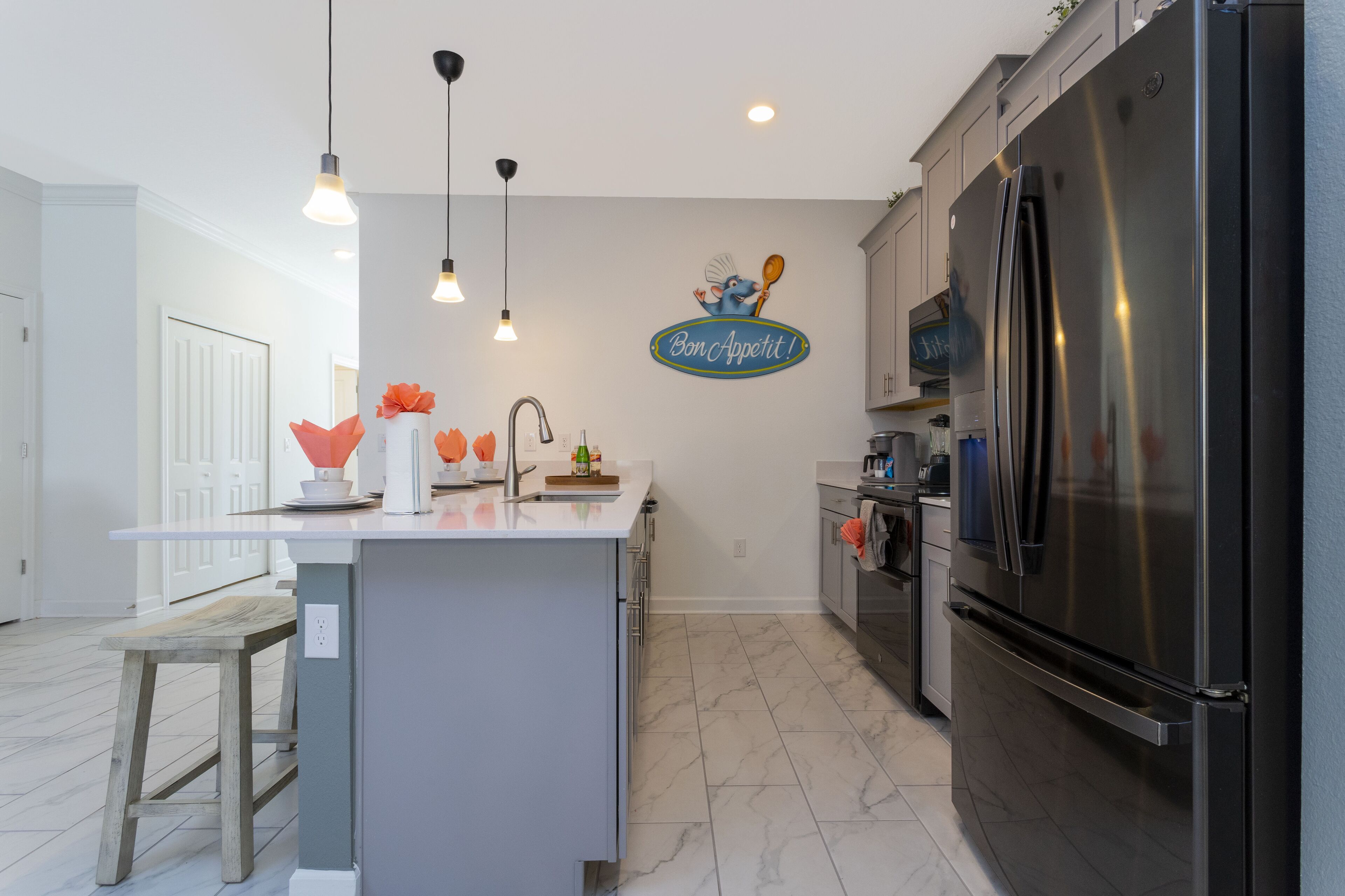 A modern kitchen with a central island, stainless steel appliances, pendant lighting, and a decorative wall sign.