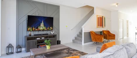 A modern living room with a flat-screen TV, a coffee table, and a sofa. There is a staircase leading to another level.
