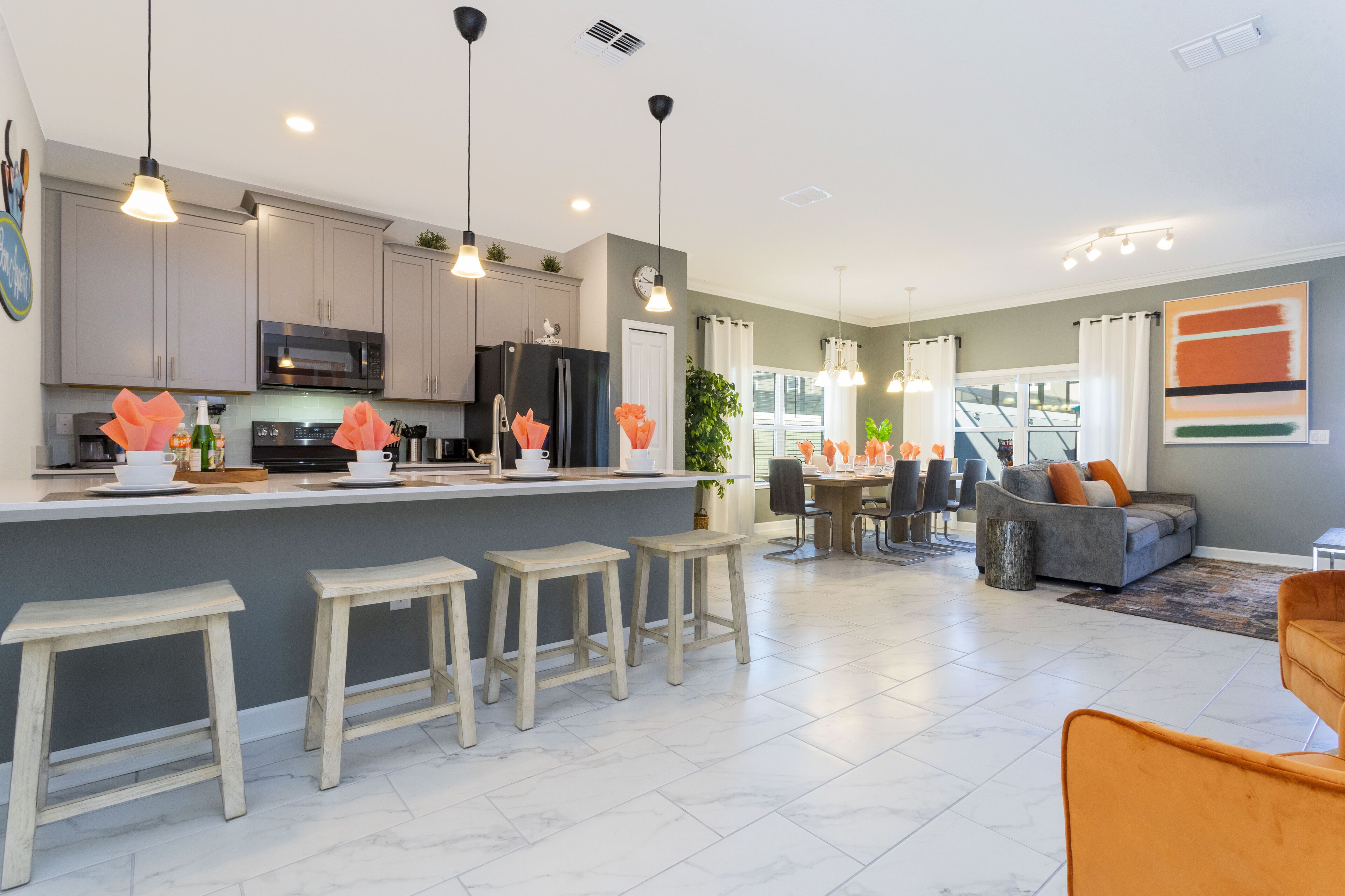 A modern kitchen with grey cabinets, a central island with stools, pendant lighting, and a dining area with a grey sofa and orange chair.