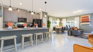 A modern kitchen with grey cabinets, a central island with stools, pendant lighting, and a dining area with a grey sofa and orange chair.