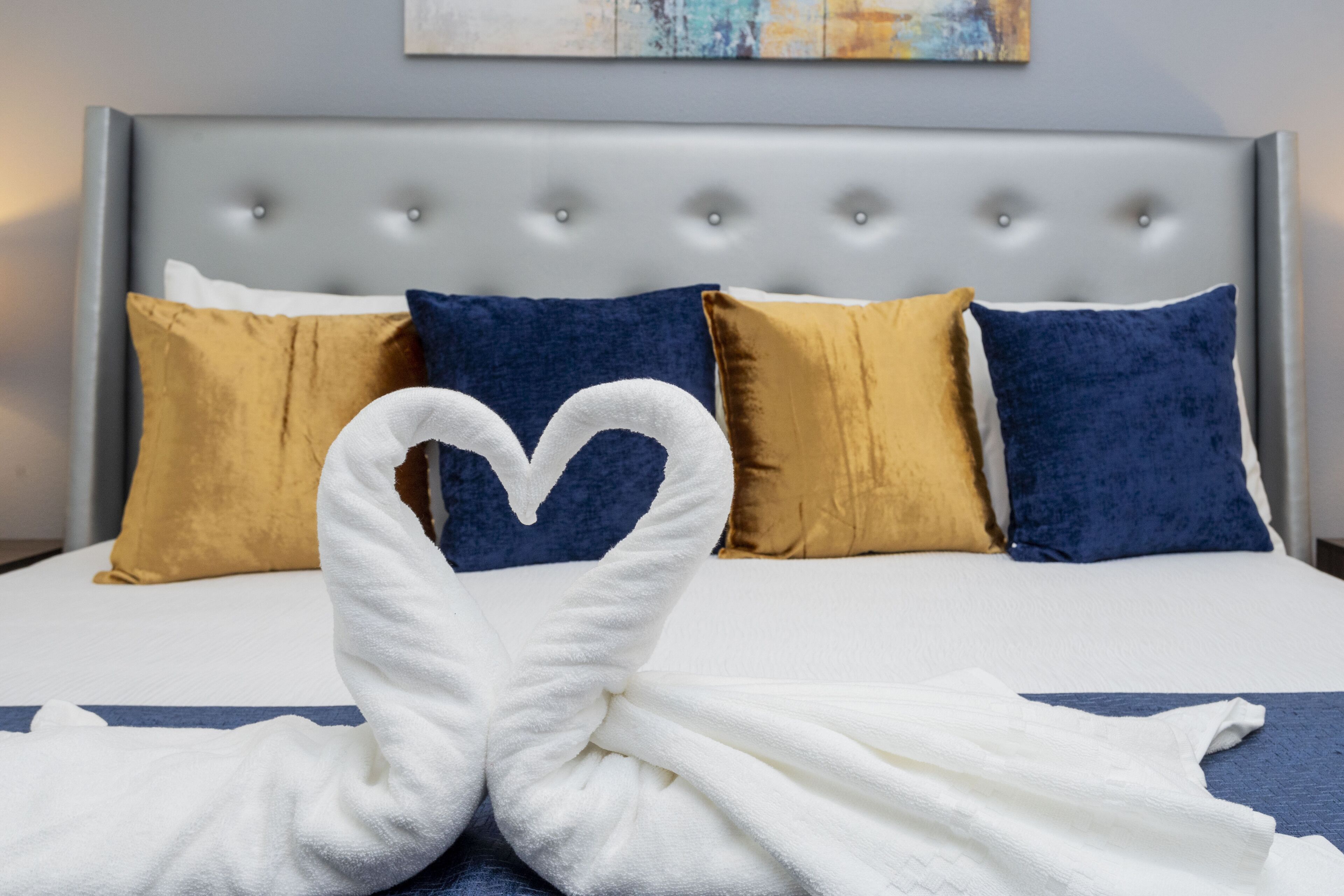 A bed with a tufted headboard, two gold and two blue pillows, and a white towel arranged in a heart shape.