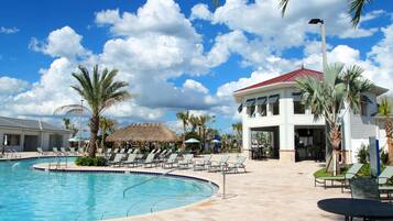 A swimming pool with lounge chairs and palm trees, a building with a red roof, and a clear blue sky.