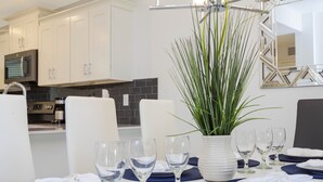 Una cocina moderna con armarios blancos, salpicadero negro y una zona de comedor con platos blancos, individuales azules y copas de vino transparentes.