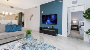 A modern living room with a teal accent wall, a flat-screen TV, a glass coffee table, and a sofa with a blue pillow.