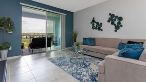 A modern living room with a beige sofa, a glass coffee table, and a blue patterned rug. There is a sliding glass door leading to a balcony with a view of the outdoors.