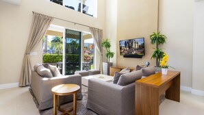 A modern living room with a grey sofa set, a wooden coffee table, and a wooden side table. There is a large window with curtains, a painting on the wall, and a potted plant. - Marvelous 4 Bd Close to Disney @ Magic Village 3153 (Kissimmee)