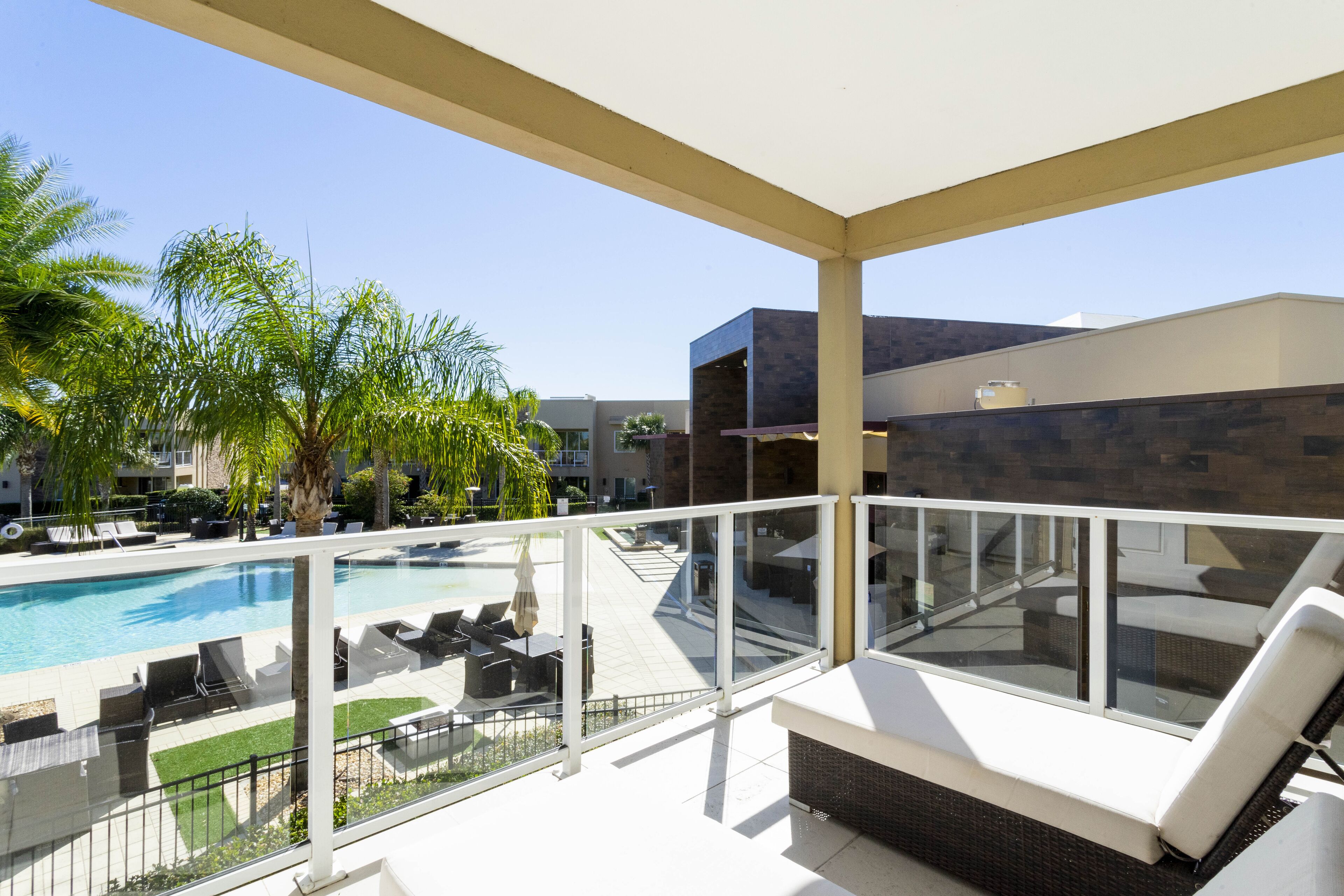 Un balcon doté d’une balustrade en verre donnant sur une piscine, des chaises longues et des palmiers.