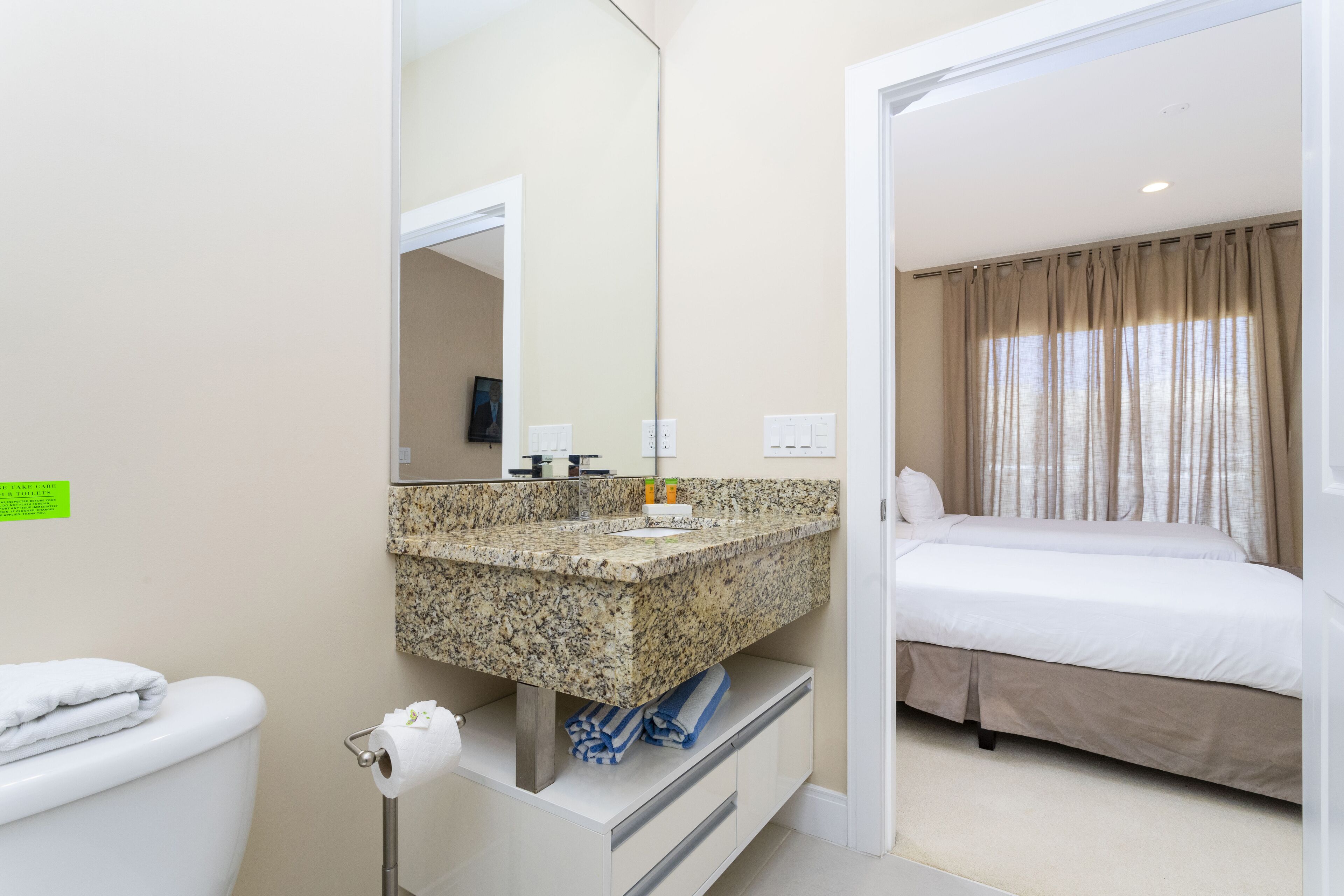 Une salle de bain d’hôtel avec un comptoir en granit, des toilettes et un lit agrémenté de rideaux beiges.