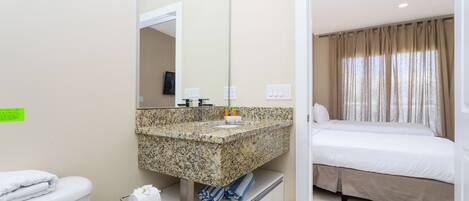 Une salle de bain d’hôtel avec un comptoir en granit, des toilettes et un lit agrémenté de rideaux beiges.