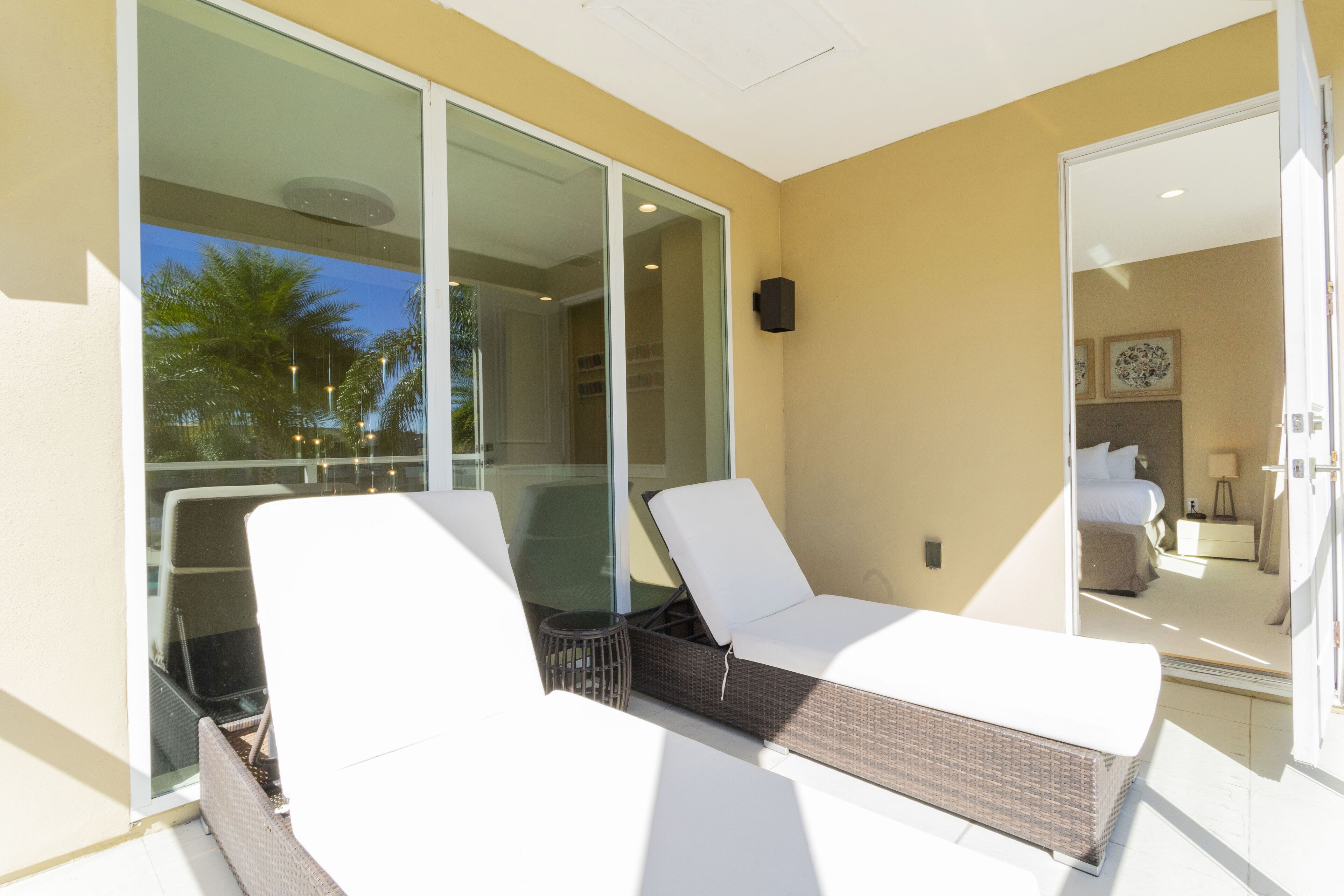 Un balcon avec des fauteuils de détente blancs et un pouf en osier, des portes vitrées donnant sur une pièce comprenant un lit et une table de chevet.