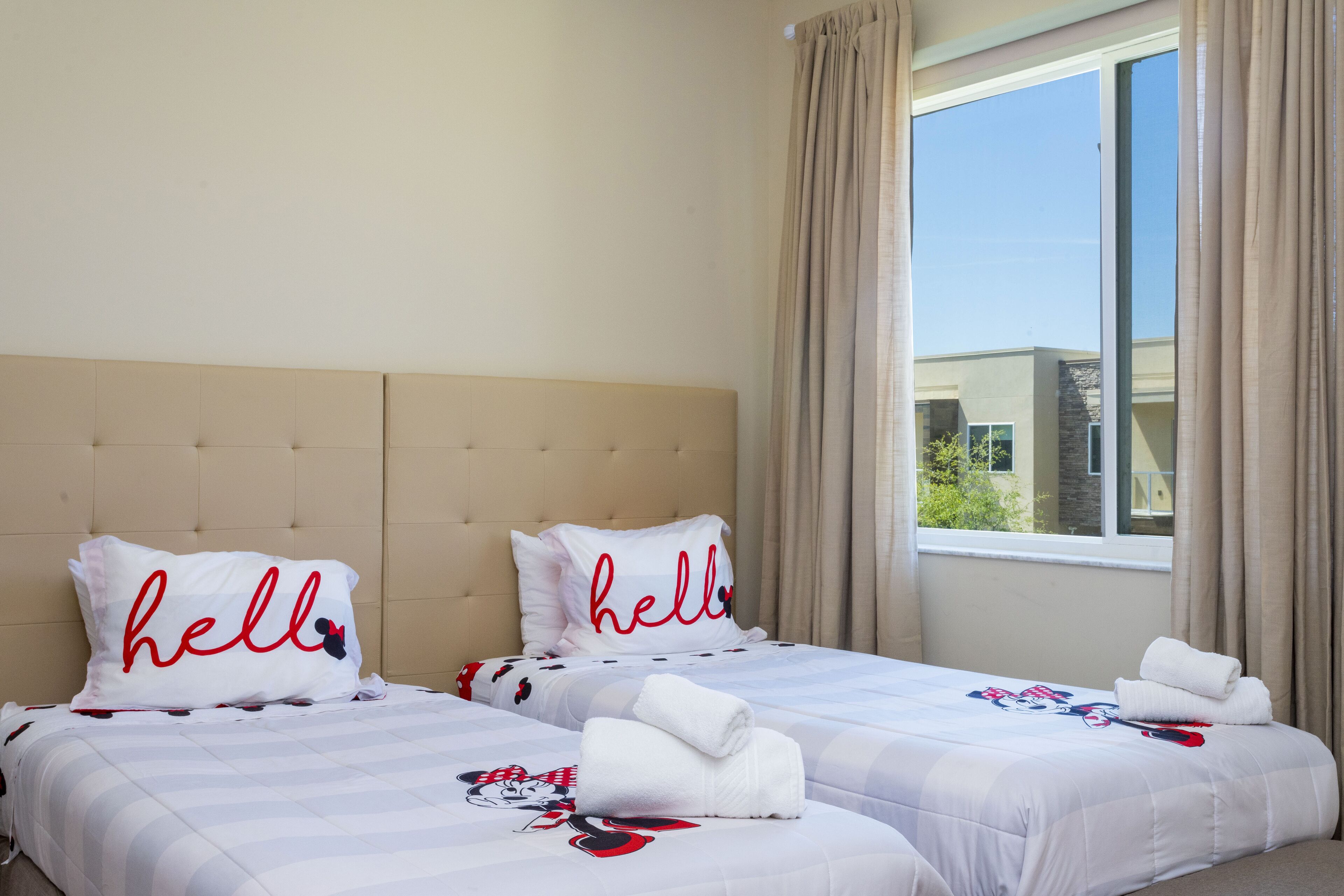 Habitación de hotel con dos camas, cabecera color crema, ventana grande con cortinas y ropa de cama temática de Mickey Mouse.