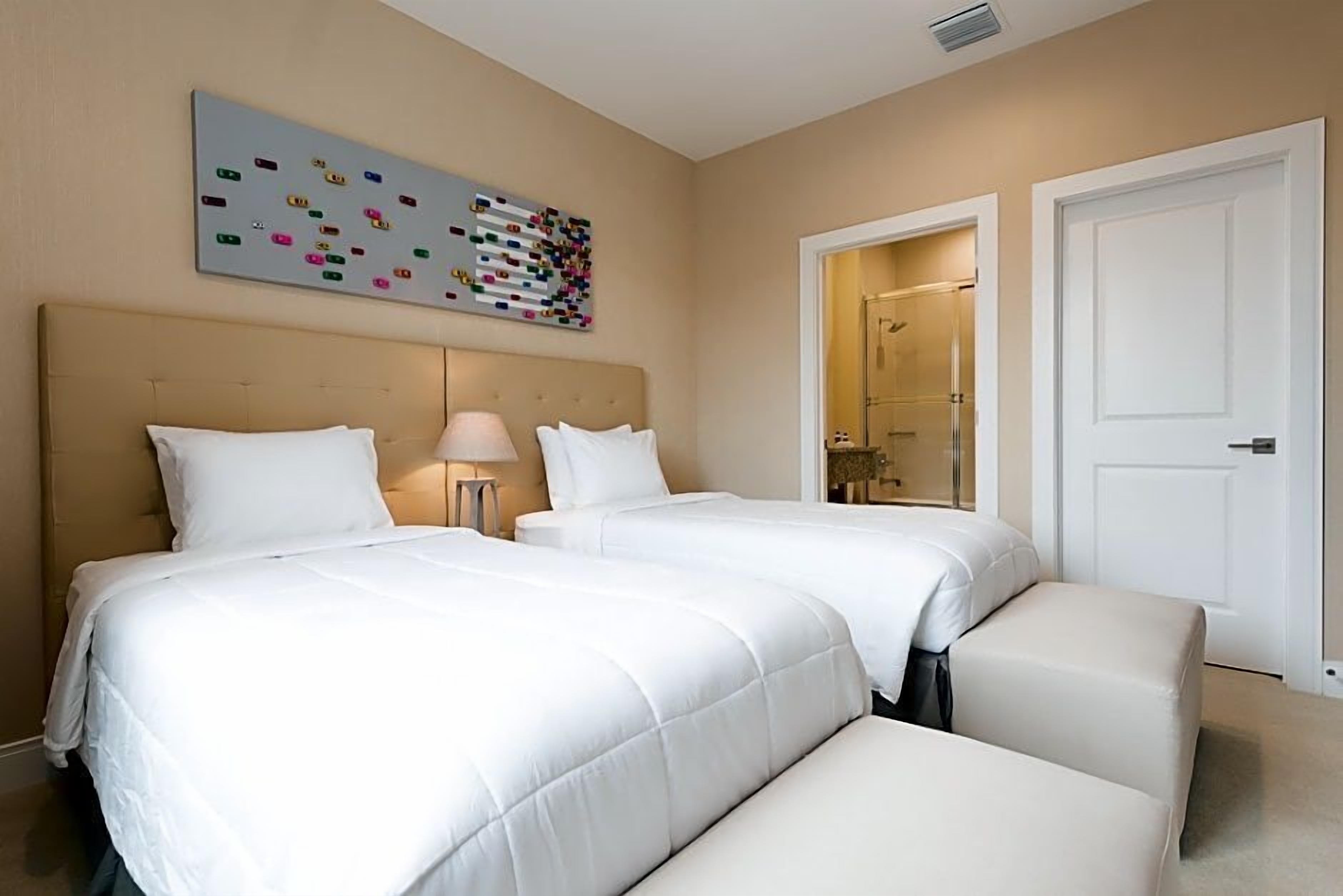 A hotel room with two beds, a bedside table with a lamp, a bathroom door, and a colorful abstract painting on the wall.