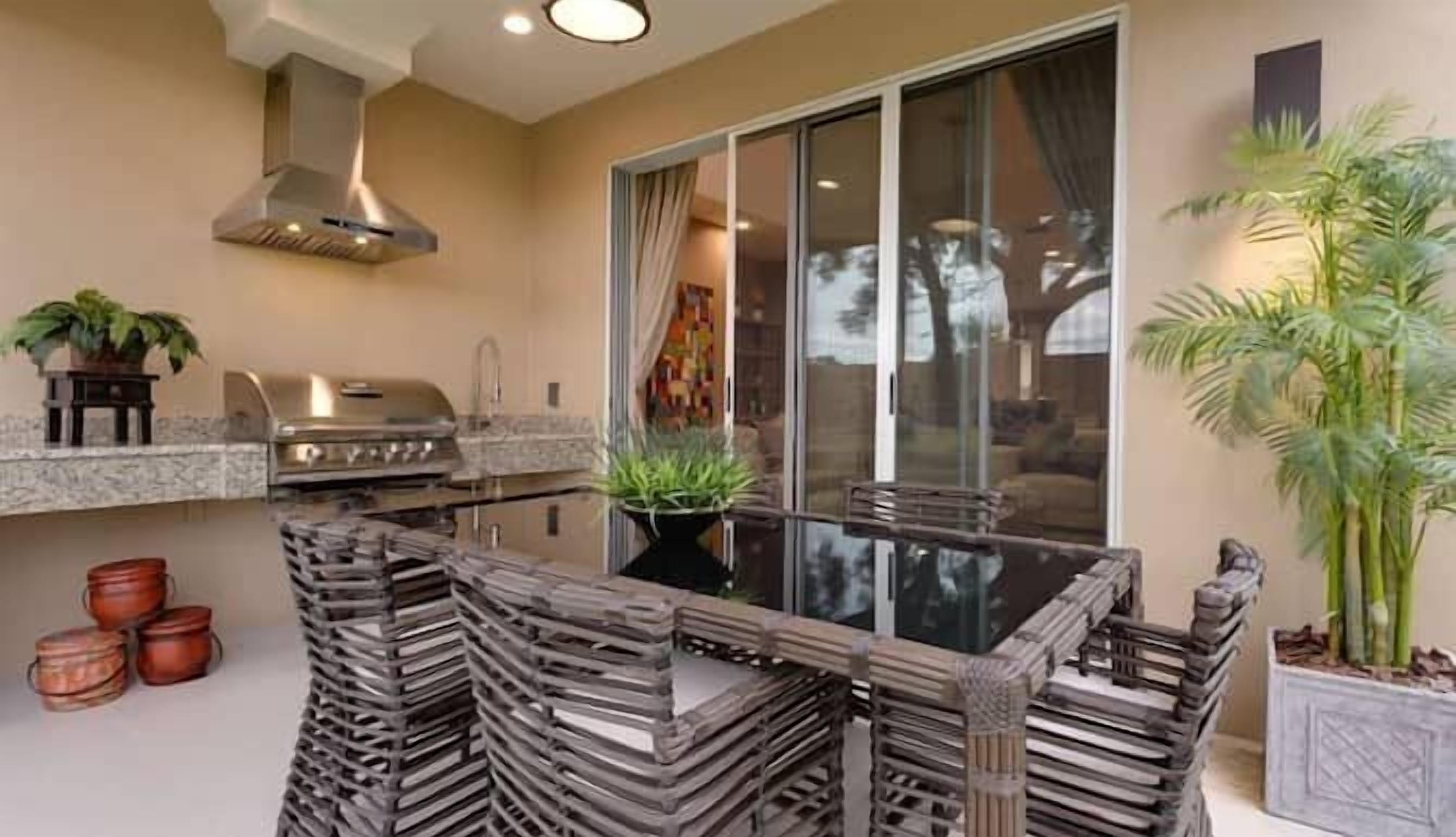 A modern outdoor kitchen with a stainless steel grill, a built-in sink, and a glass-top dining table surrounded by wooden chairs.