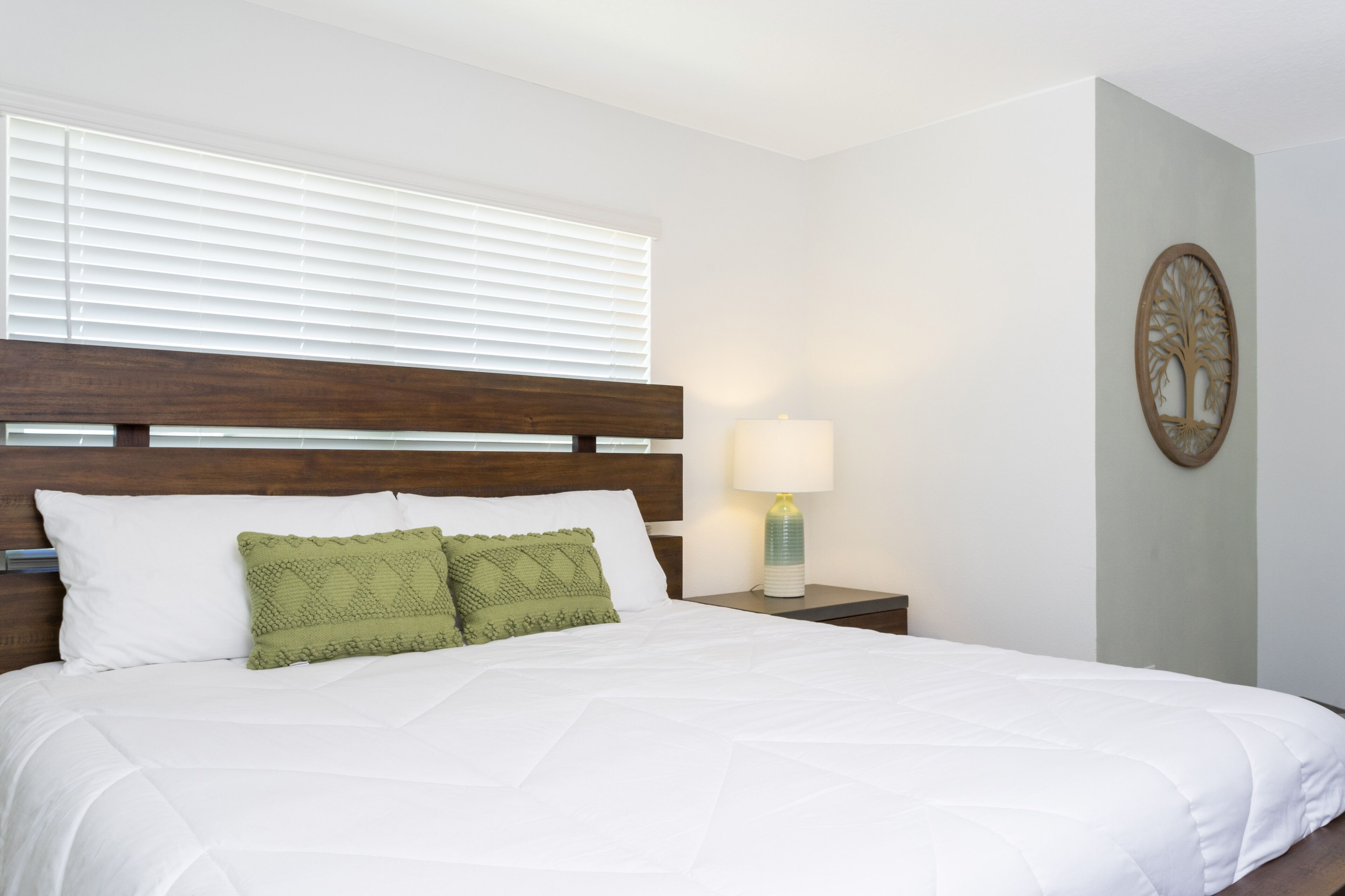 A neatly made bed with white bedding and two green pillows, a wooden headboard, a bedside lamp, and a wall-mounted tree decoration.