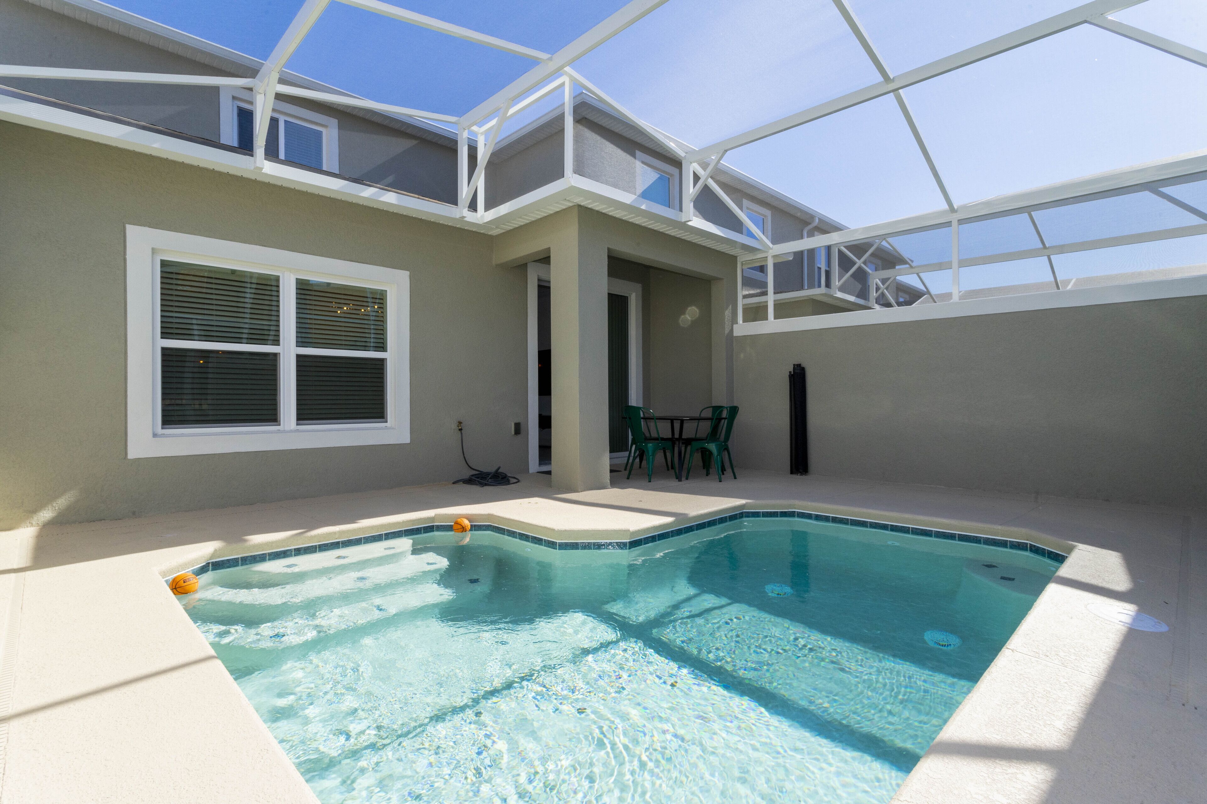 A swimming pool with a glass ceiling, a window with blinds, and a patio area with chairs.