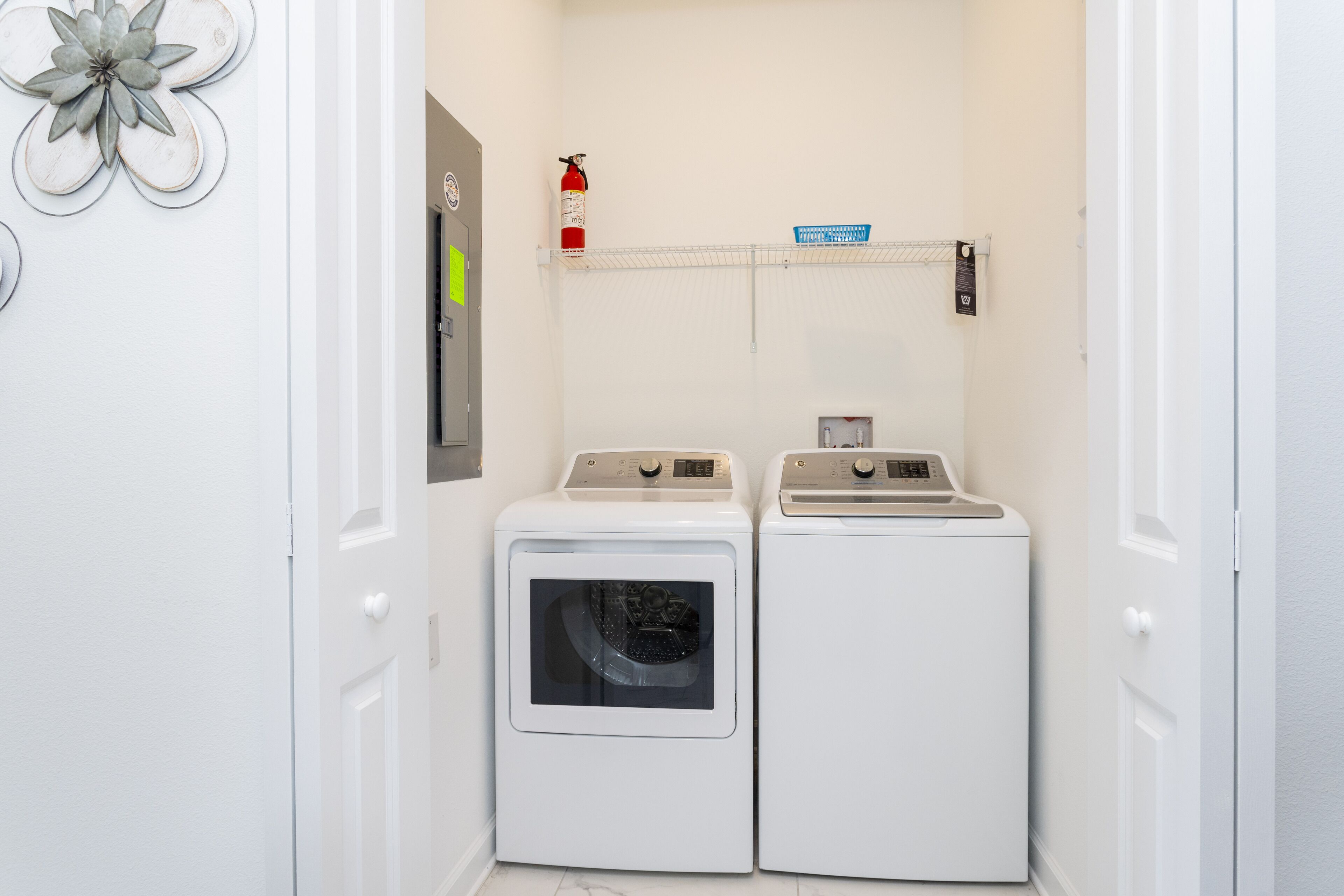 A laundry room with two white front-loading washing machines, a fire extinguisher, and a shelf with a blue basket.
