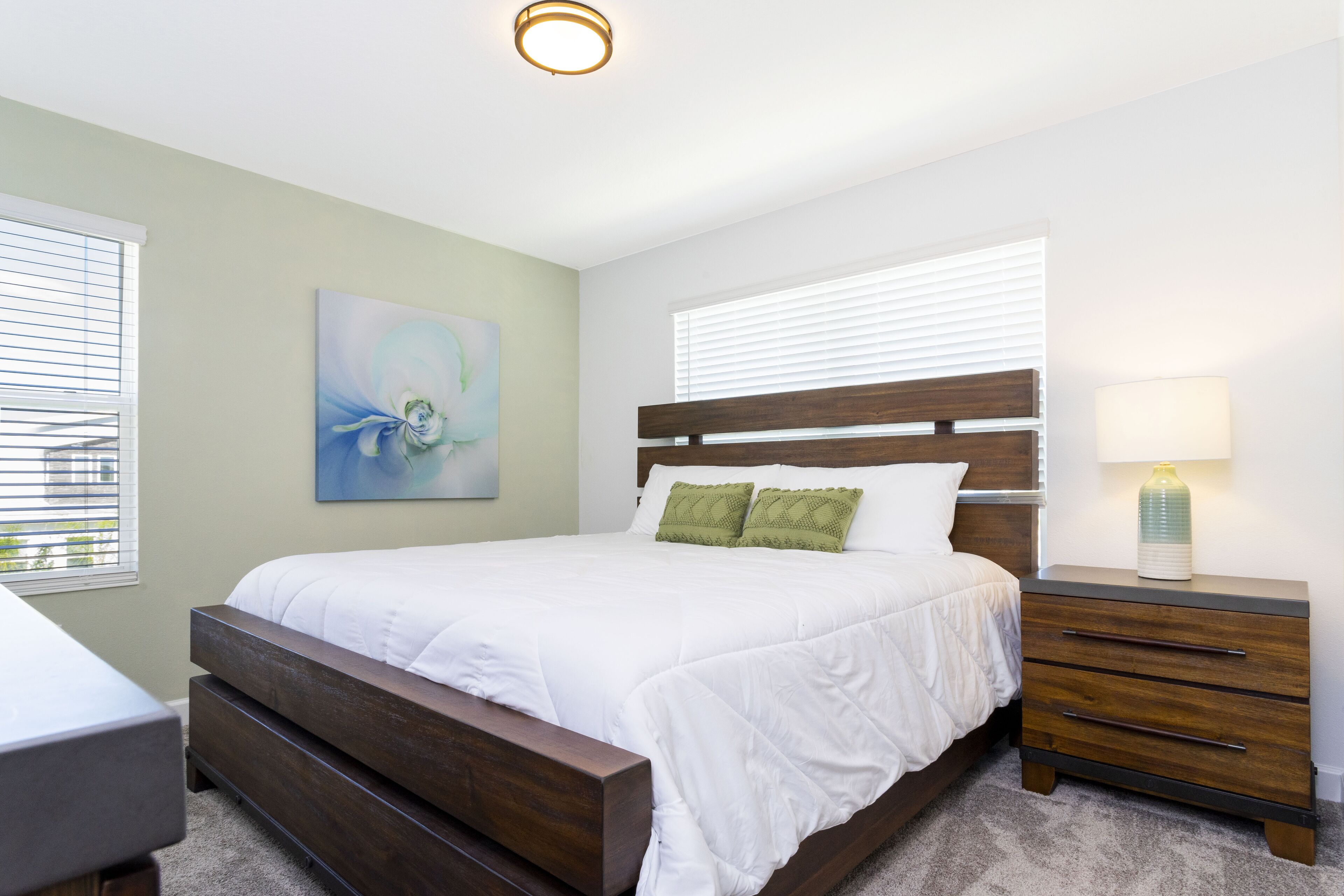 Quarto com cama de madeira, mesa de cabeceira com abajur e um quadro na parede.