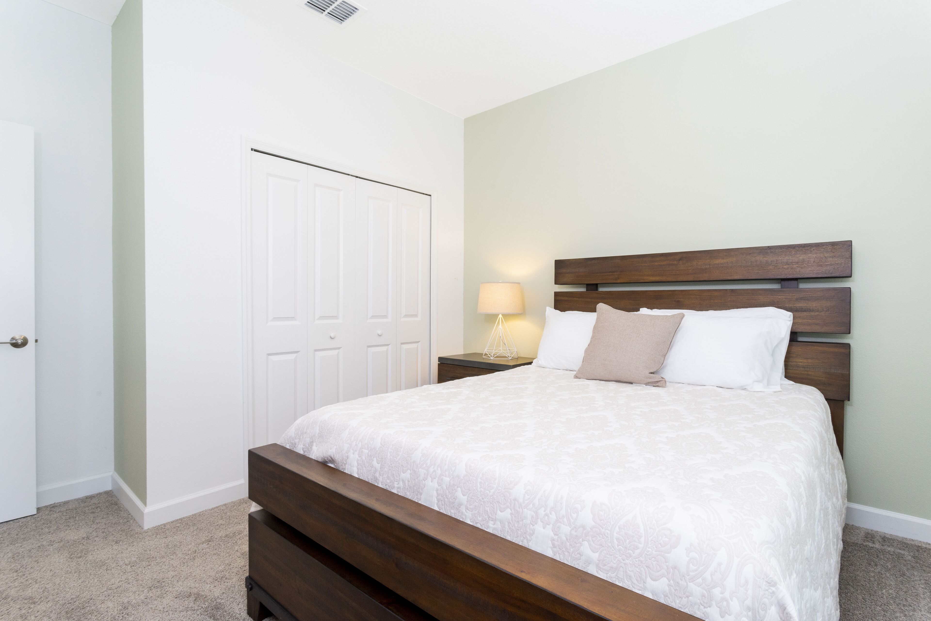 Een slaapkamer met een houten bed, witte beddengoed, een nachtkastje met een lamp en een witte deur.