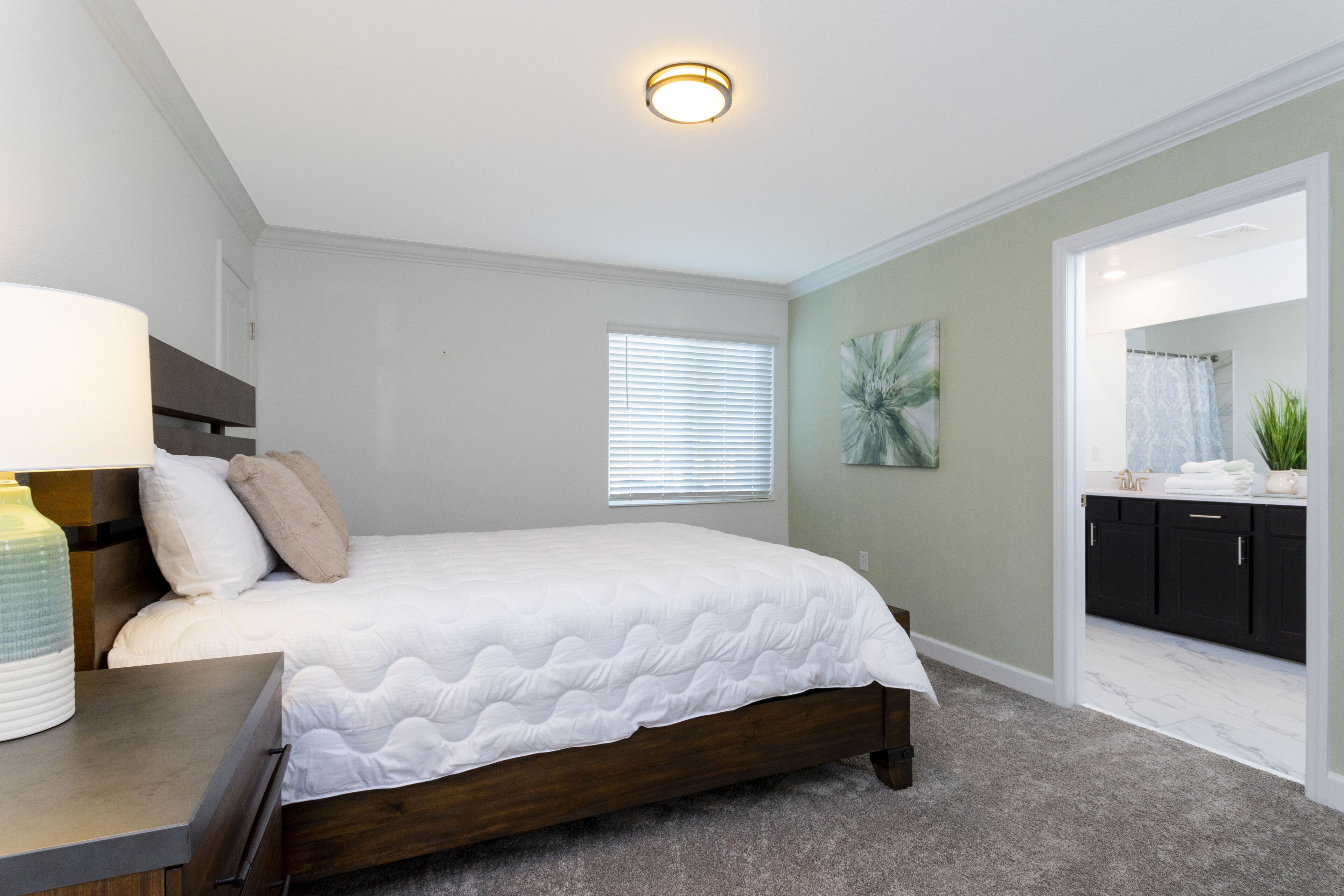 A bedroom with a bed, bedside table, lamp, window, and a view into the bathroom.