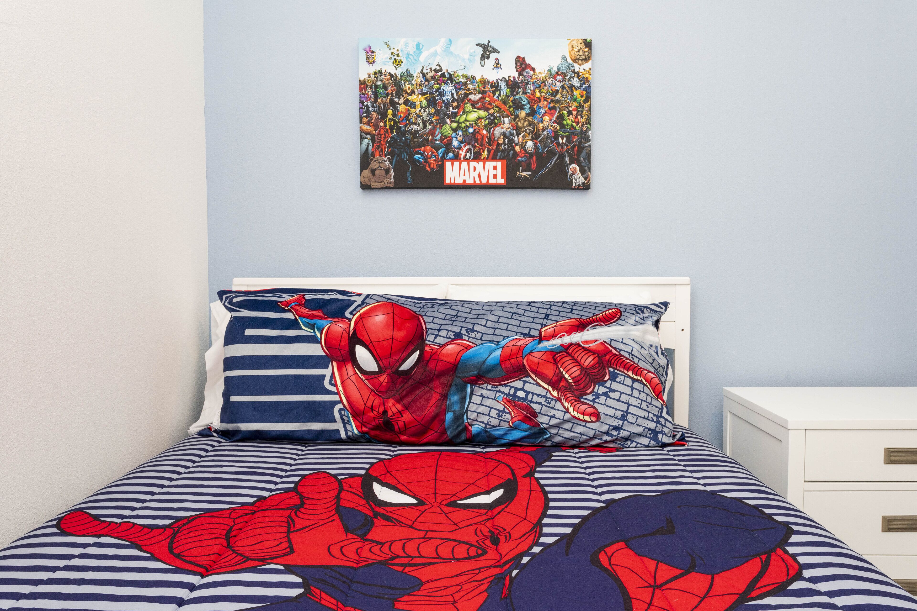 A bedroom with a bed featuring Spider-Man themed bedding, a wall-mounted poster of Marvel characters, and a white nightstand with drawers.
