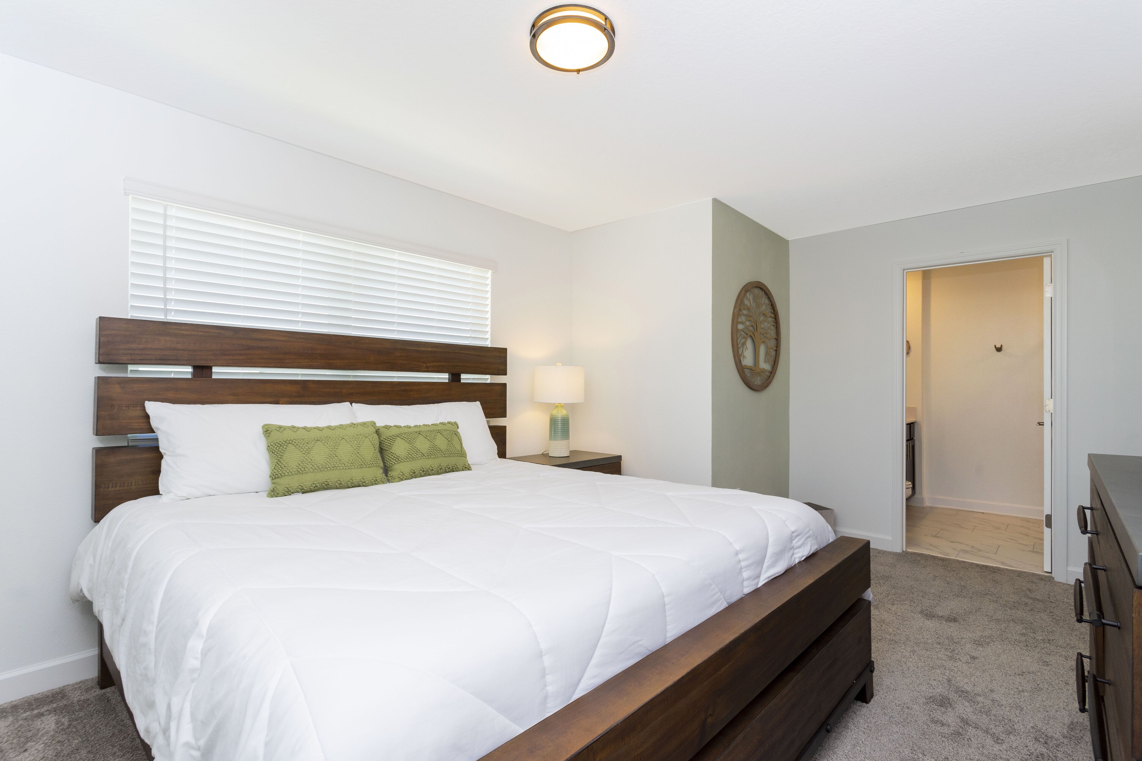 A bedroom with a wooden bed, white bedding, a nightstand with a lamp, and a door leading to another room.