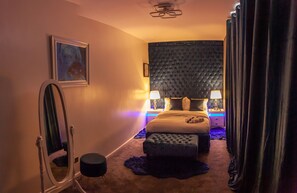 Ein Schlafzimmer mit einem großen Bett, zwei Nachttischen mit Lampen, einer Kopfteil, einem Spiegel und einem gerahmten Bild an der Wand.