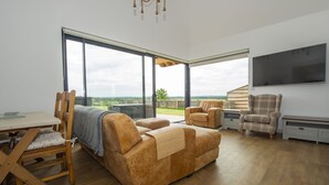 A spacious living area with a large sliding glass door leading to a balcony, a wooden dining table with chairs, a sofa, and two armchairs. - Owl Lodge (Wedmore)
