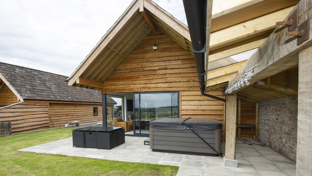A wooden cabin with a patio area featuring a hot tub, outdoor seating, and a stone wall. - Owl Lodge (Wedmore)