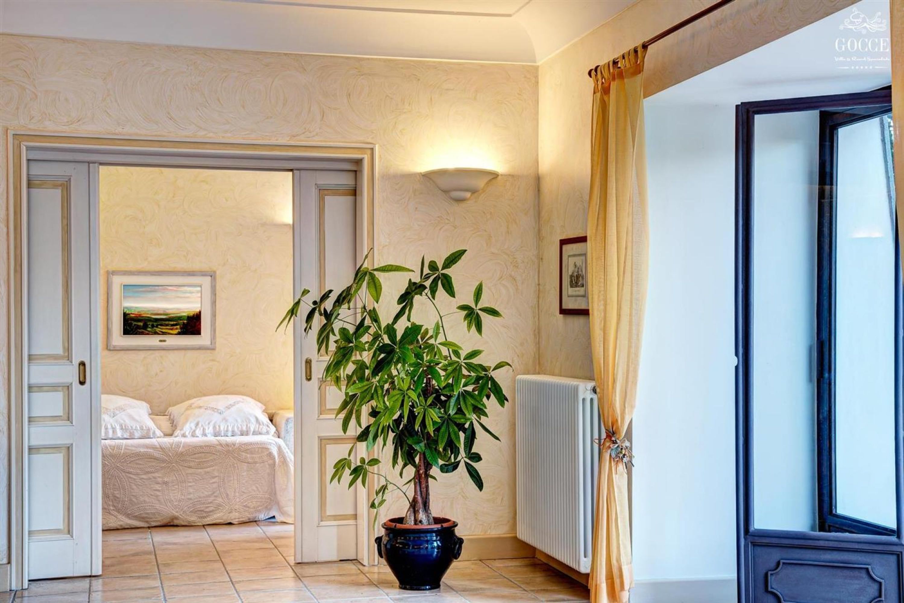 A hotel room with a bed, a potted plant, a radiator, and a framed picture on the wall.