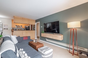 Una sala de estar moderna con un sofá gris, un reposapiés estampado, una mesa de centro y un televisor de pantalla plana montado en la pared.