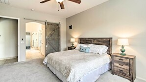 Quarto com cama, cabeceira de madeira, duas mesas de cabeceira com luminárias, um ventilador de teto e uma porta que dá para outro cômodo.