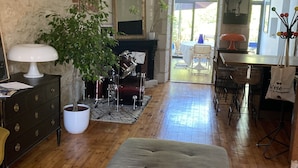 A living room with a wooden floor, a grey ottoman, a white lamp, a potted plant, a drum set, and a fireplace. - Charming town house with pool and garden (Périgueux)