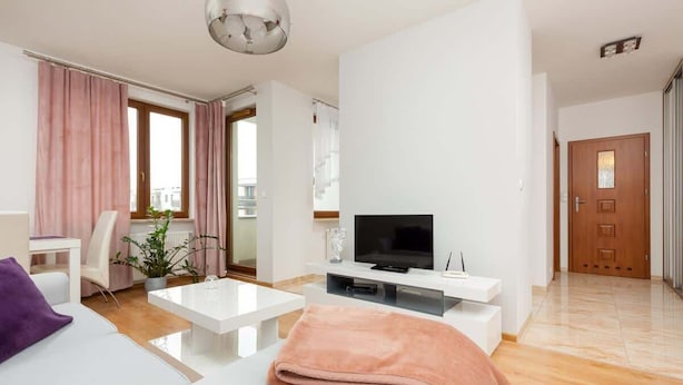 Una sala moderna con sofá, una mesa de centro y un mueble para la televisión. Se ve una ventana con cortinas, una planta y una puerta al fondo.