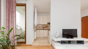Un apartamento moderno con sala de estar, televisor, planta y cocina al fondo.