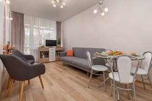 A modern living room with a sofa, dining table, and chairs, a TV, and a window with curtains. - Tasteful Apartment | Balcony | 750 m to the beach (Łukęcin)