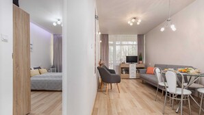 A modern living room with a sofa, dining table, and chairs, a TV, and a bed in the background. - Tasteful Apartment | Balcony | 750 m to the beach (Łukęcin)