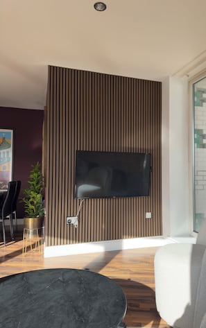 A modern living room with a flat-screen TV mounted on a wooden slat wall, a black round coffee table, a grey sofa, and a potted plant.