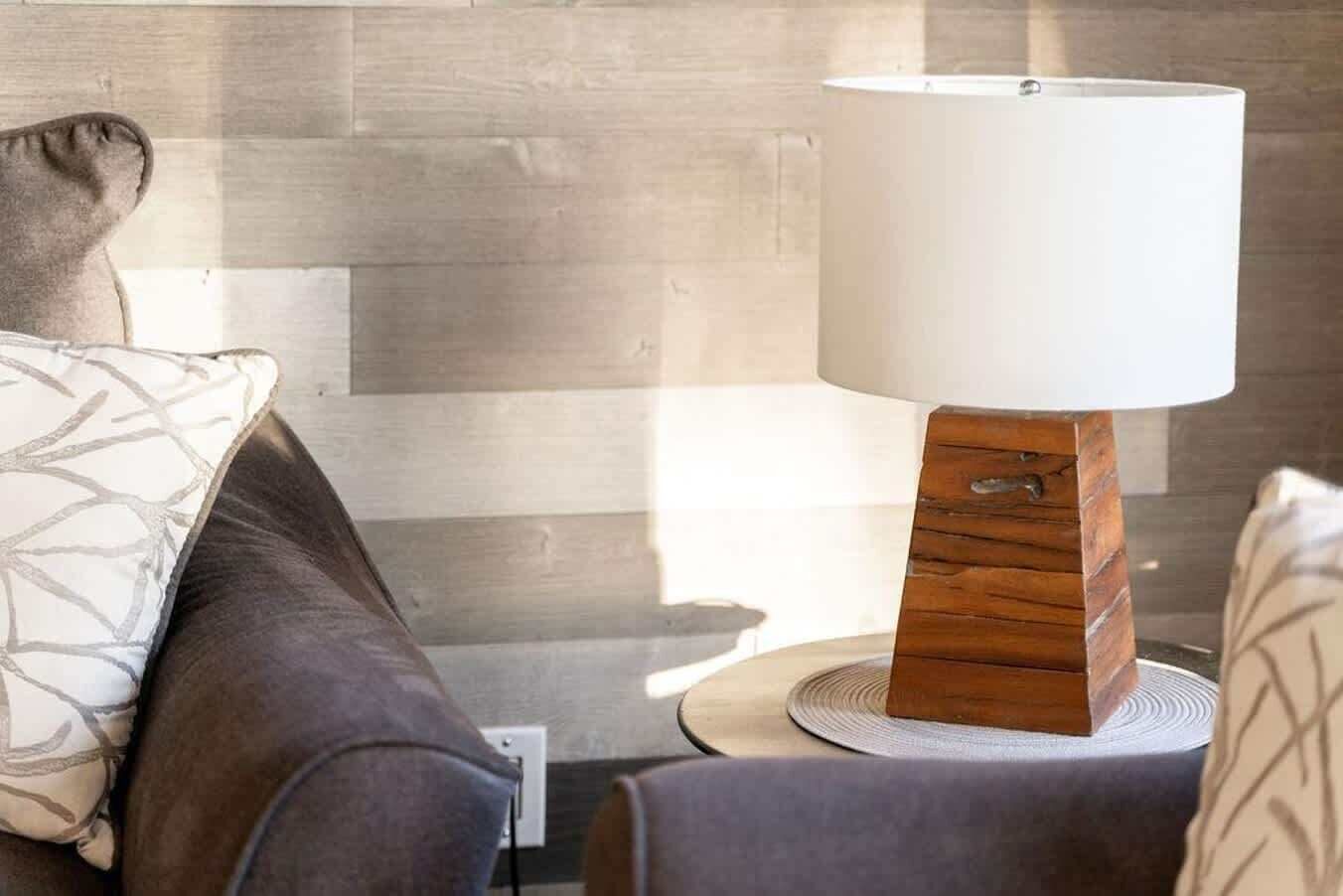 Una sala moderna con una lámpara de pie de madera sobre una mesa redonda, un sofá gris con cojines estampados y una pared lisa al fondo.