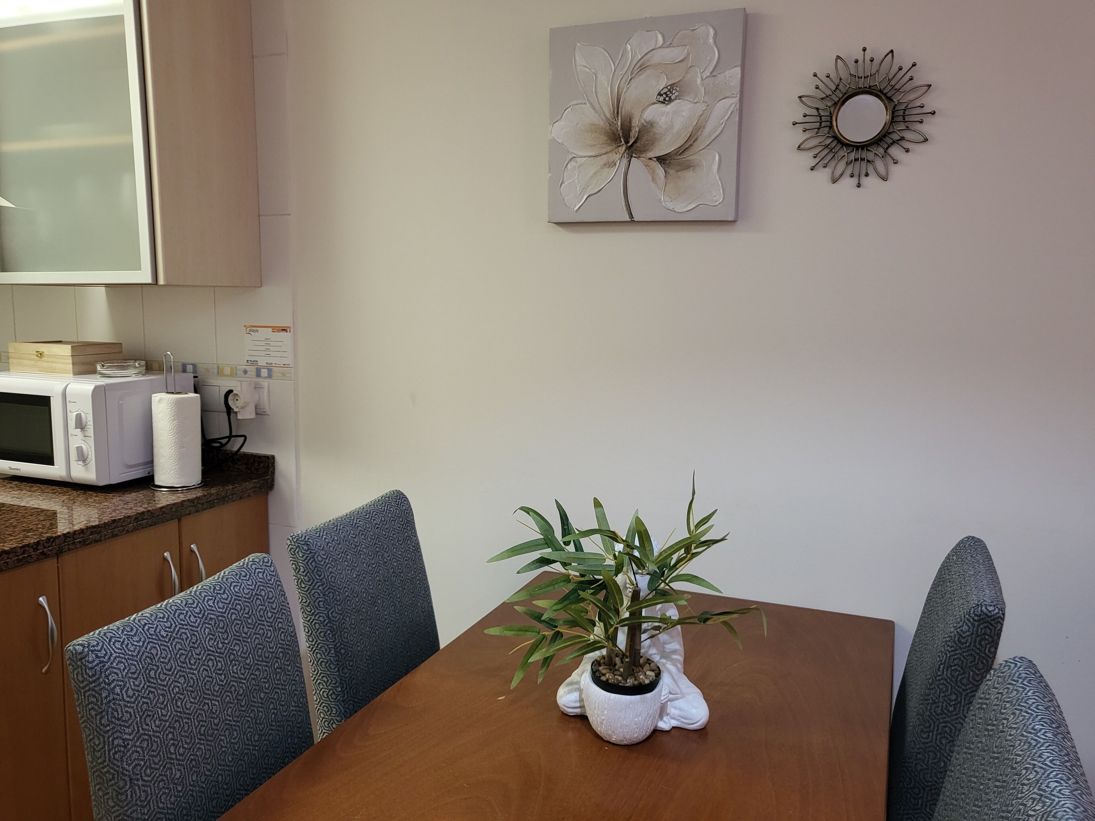 Un comedor con una mesa de madera, sillas estampadas, una planta en maceta, un microondas y un cuadro de flores colgado en la pared.