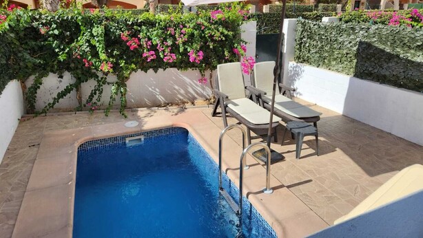 A swimming pool with a tiled edge, surrounded by a white wall with green climbing plants and pink flowers. There are two lounge chairs and a large umbrella providing shade. - Relaxing essence bass with private pool and patio! (Vera)