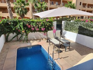 A swimming pool with a tiled edge, surrounded by a white wall with green climbing plants and pink flowers. There are two lounge chairs and a large umbrella providing shade. - Relaxing essence bass with private pool and patio! (Vera)