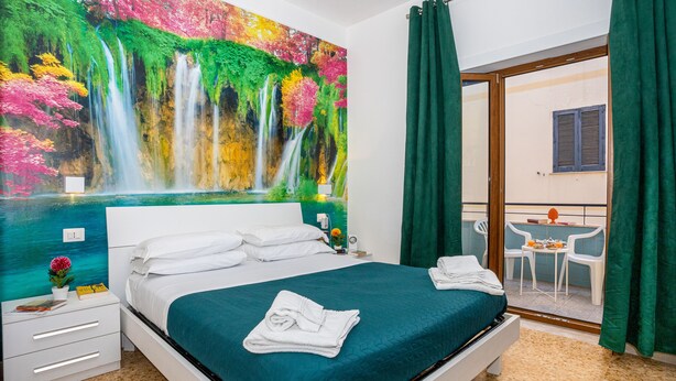 Hotellihuone, jossa on suuri sänky, yöpöytä, pieni pöytä ja tuoli, näkymä parvekkeelle sekä maalaus vesiputouksesta.