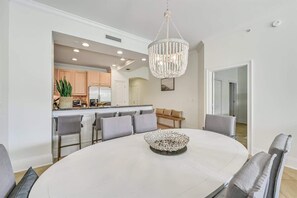 A dining area with a round table, grey upholstered chairs, a chandelier, and a kitchen in the background. - Steps to Beach,Gulf Front Pool,Hot Tub-Vitamin Sea (Santa Rosa Beach)