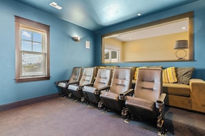 A room with four reclining chairs, a window with blinds, a mirror, a lamp, and a couch with cushions.