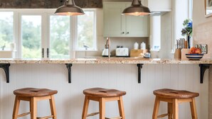 A kitchen with a granite countertop, three wooden stools, hanging pendant lights, and a view of the outdoors through large windows.