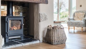 A wood-burning stove with a stone surround, a wicker basket of firewood, and a patterned armchair in a room with wooden flooring and a large window.