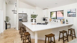 A modern kitchen with white cabinetry, a large island with stools, a stainless steel refrigerator, and a window with a view. - Upscale Red Rock Retreat- Spa, Views, Pet Friendly (Sedona)