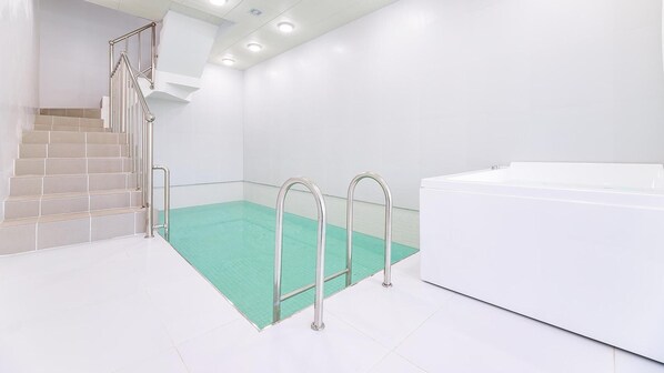 Una piscina interior moderna con una barrera de cristal, una escalera con barandillas de metal y una bañera jacuzzi blanca.