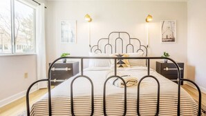 A modern bedroom with a metal bed frame, two bedside tables, a large window, and wall art.